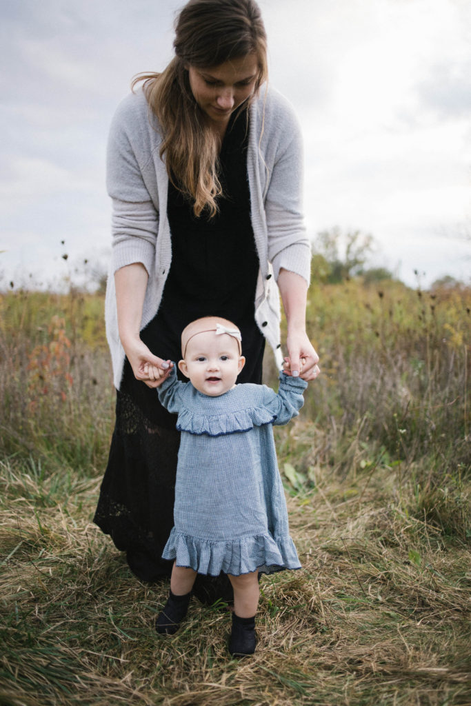 Large family sessions with Elle Baker Photography near Chicago. A mom helps a young baby girl in a field learning how to walk 