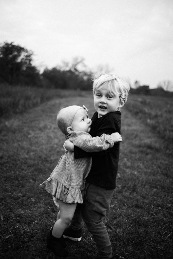 Large family sessions with Elle Baker Photography near Chicago. A young boy carries his baby sister down a pathway. Both wearing clothing from Zara during a photosession