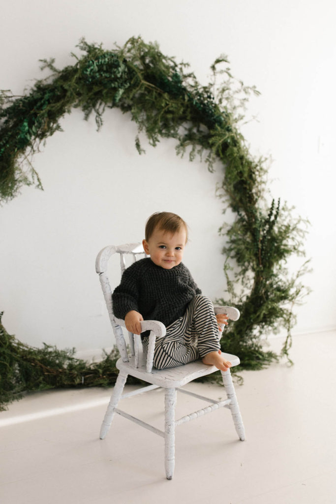Homer Glen, IL Holiday Mini Sessions, Photo by Elle Baker Photography, boy sitting on a white chair with a large wreath during Christmas session