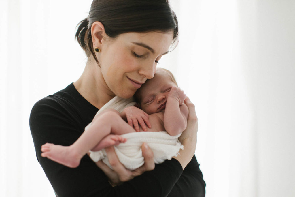 Smiling mother holds her newborn baby boy in front of a window and white curtains