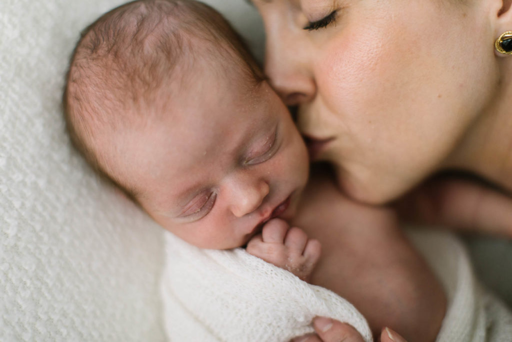 Mother kissing newborn baby on the cheek, newborn wearing a white wrap on a white backdrop during simple session
