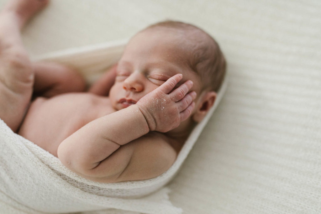 newborn baby's hand rests on his cheek during a newborn session, baby wrapped in ivory organic wrap with torn edges