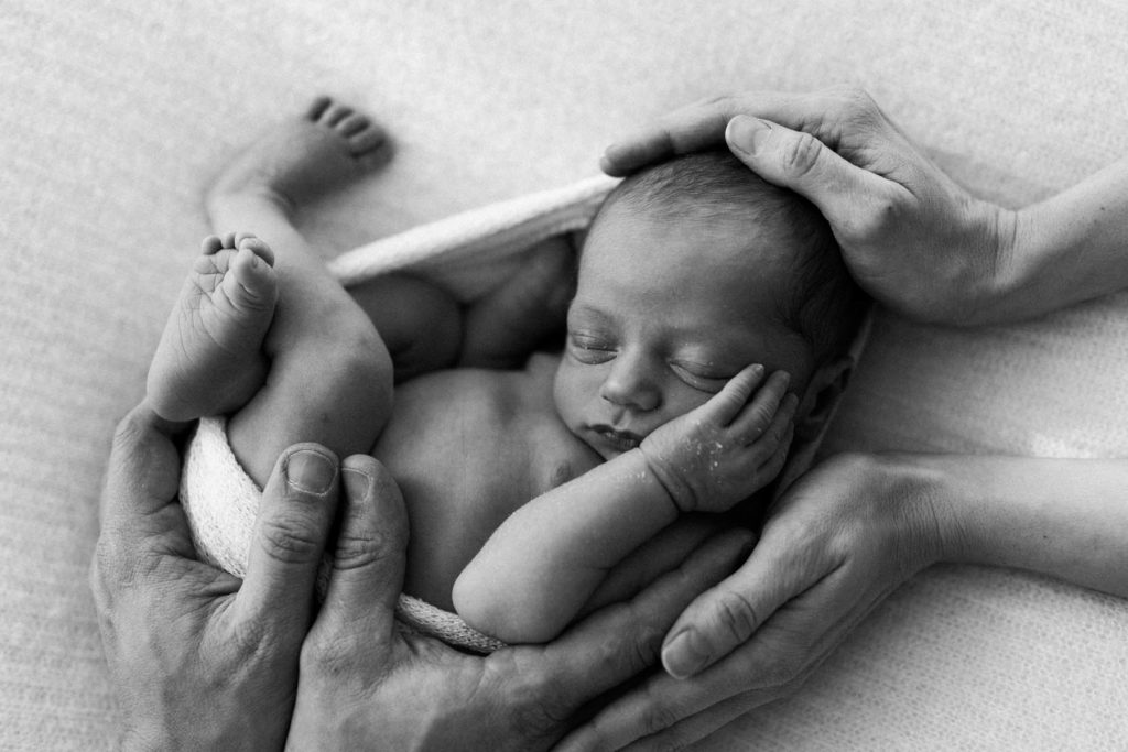 black and white image of mother and father's hands wrapped around their newborn baby 