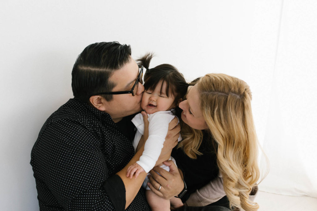 family of three, mom and dad kissing baby girl on the cheek at the same time