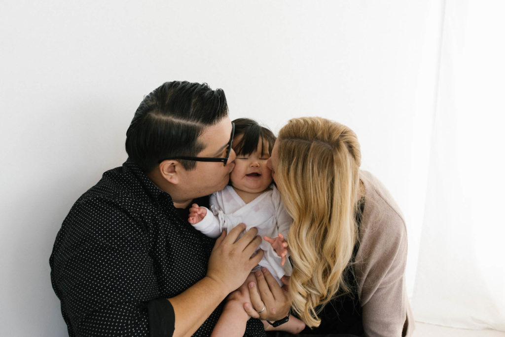 mom and dad kissing baby girl on the cheek at the same time