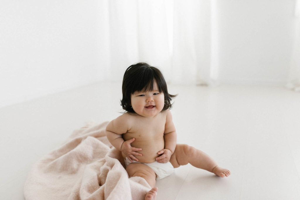baby girl laughs while holding her tummy, she has dark hair and is wearing a diaper