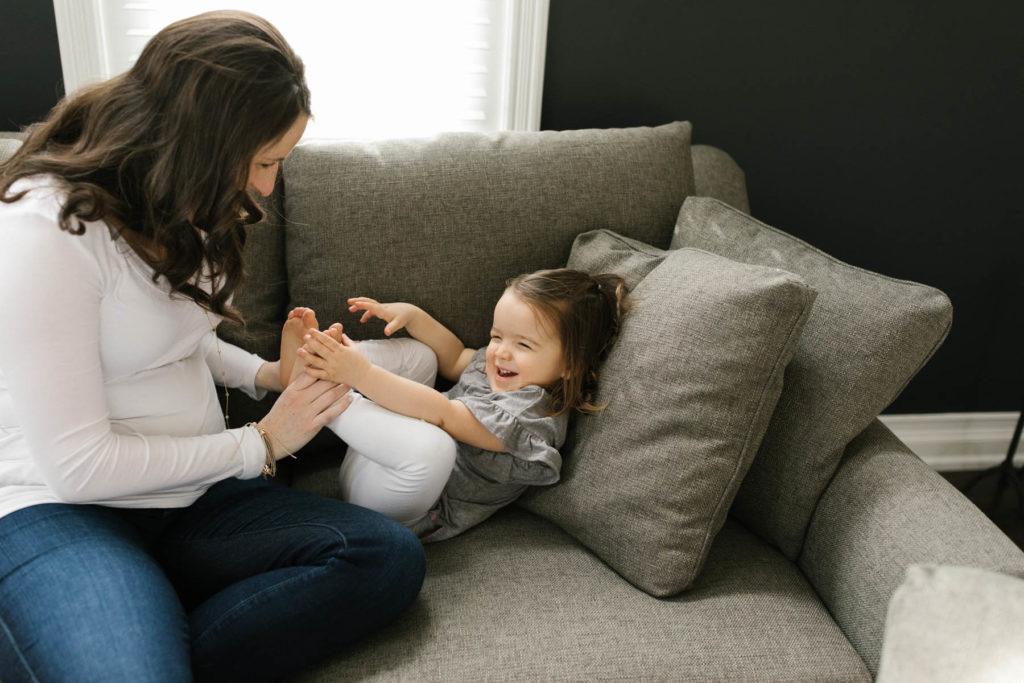 Mom tickles her daughters toes on a gray couch during lifestyle session 