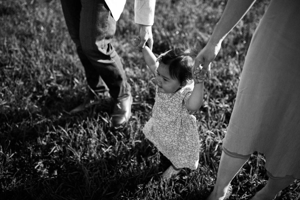 Mom and dad holding little girls hands, helping a toddler walk during family session