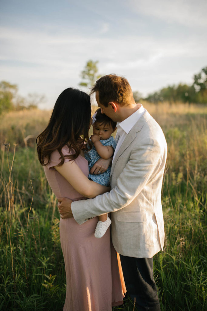 family of three cuddling baby girl who is sucking her thumb, mom in a rose colored dress, dad in a sports coat