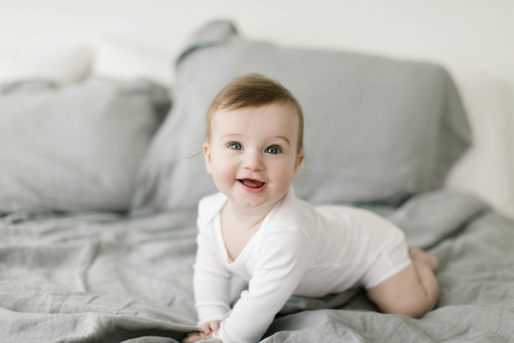 baby smiles and looks past the camera on a bed with gray linen covers 