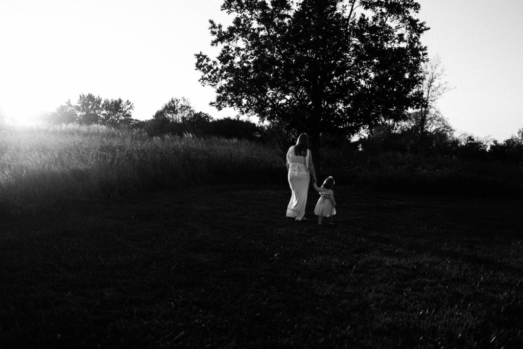 Maternity and Family session near Frankfort Illinois Mom and daughter holding hands walking outside during sunset session. Mom is wearing a white gown and showing her baby bump