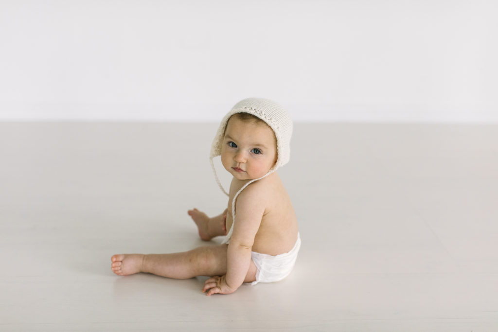 Laurie photographs baby led sitter session with a little girl in a bonnet in her white studio