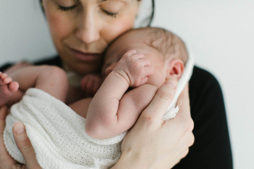 close up photo of mother holding newborn baby wrapped in a neutral wrap against a white wall