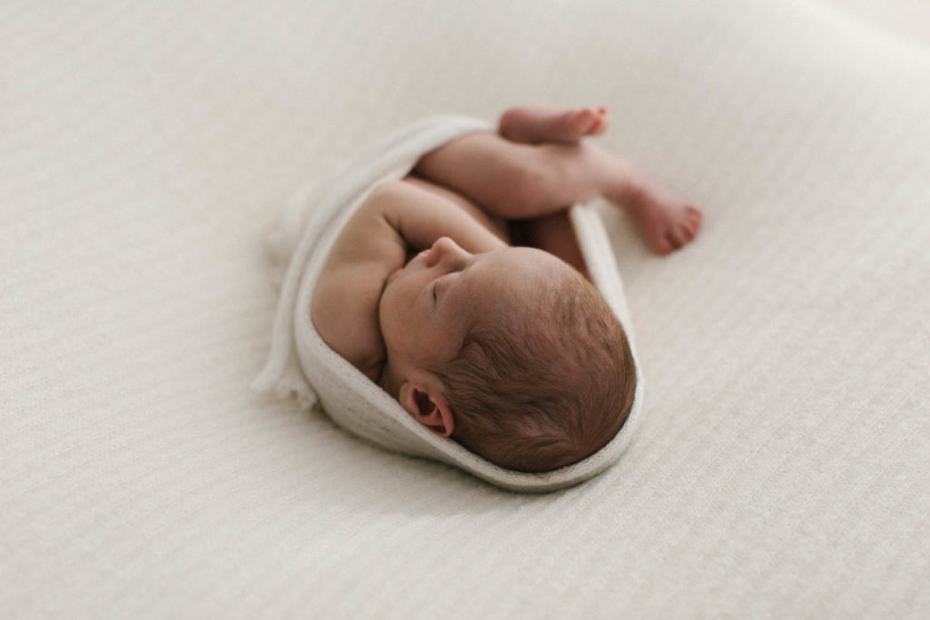 newborn baby curled in a fetal position on a white blanket and wrapped in a torn edge ivory wrap