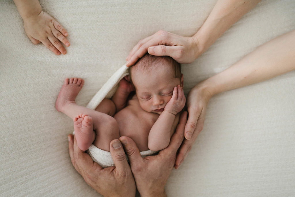 mother and father wrapping their hands around their newborn baby and small sibling's hand reaches into the frame