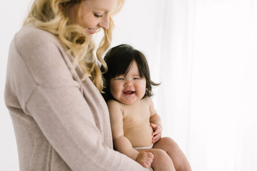 baby girl with dark black hair smiles as her blonde mom holds her