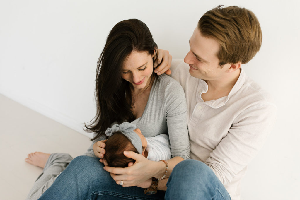 family of three with newborn baby, woman with dark hair and husband smiling at her