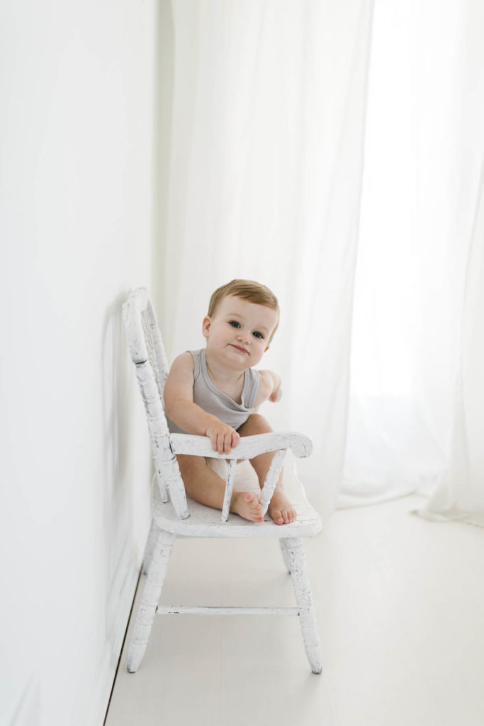 cute baby sitting on white chair in a neutral and white studio 